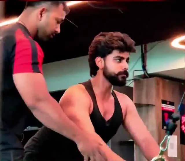 A WTF coach in a red and black team tee guiding a member through a cable exercise.