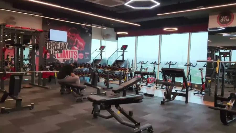 A wide WTF gym floor with benches, dumbbell racks and a row of bikes along a glass wall